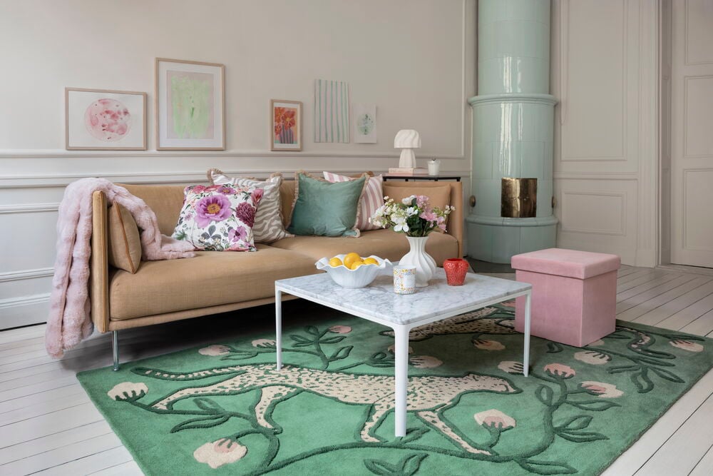 Grön ullmatta med gepard och rosa blommor, brun soffa med mönstrade kuddar, rosa pläd, marmorbord, rosa ottoman.