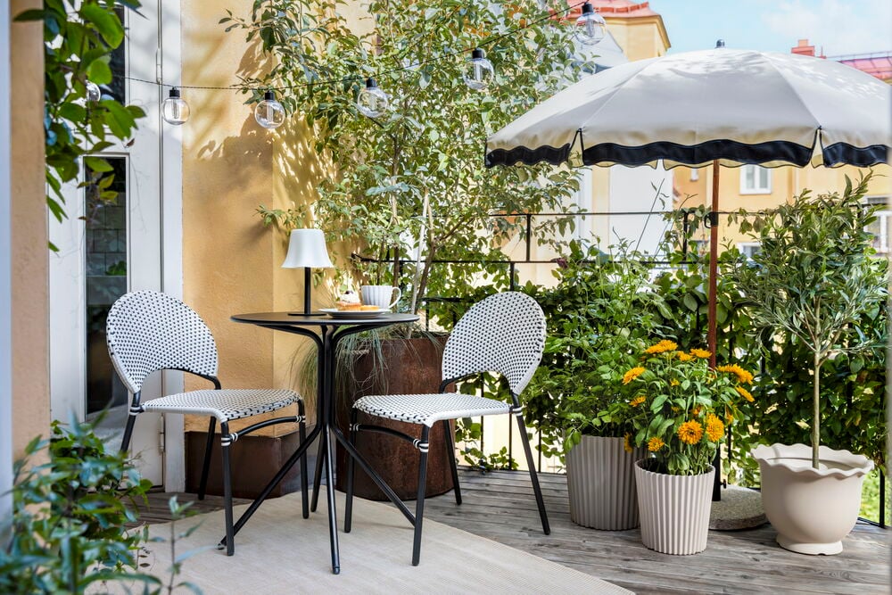 Cafébord Bistro Ø60 cm Svart Stål, två svartvita flätade stolar plus ett vitt uteplats-parasoll.