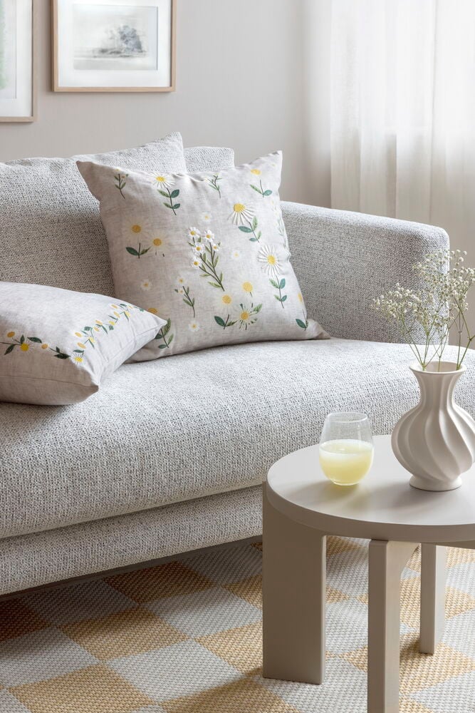 Putetrekk Maya Natur Polyester med tusenfryd-broderi og et matchende putetrekk på en lys grå teksturert sofa.