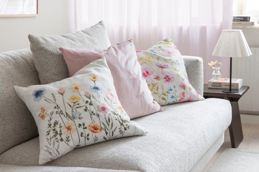 Vanha pinkki polyesterisametti Butterfly tyynynpäällinen ja kaksi valkoista kukallista tyynynpäällistä harmaalla sohvalla.