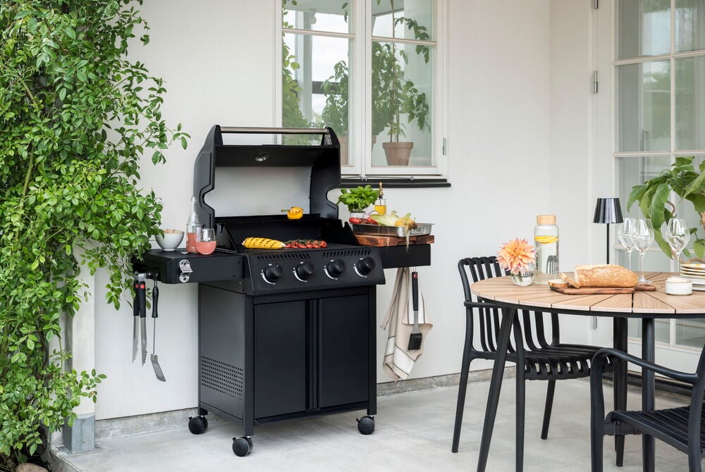 Musta Kaasugrilli Titan pyörillä, grillattua ruokaa, ruoanlaittovälineitä ja juomia sivuhyllyillä.