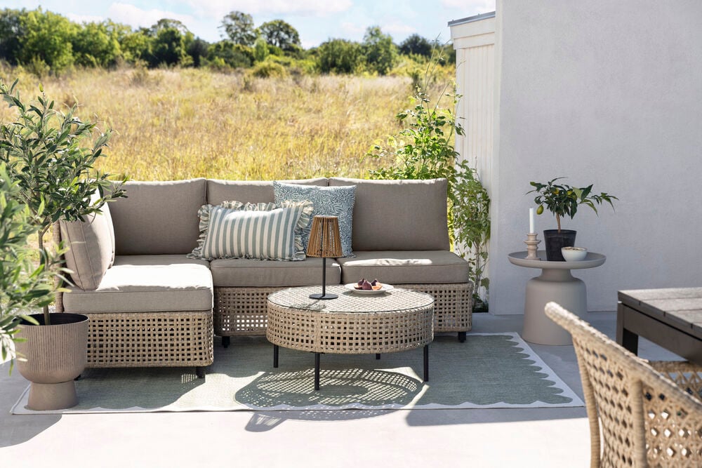 Beiger runder Beistelltisch Tunis Beige, mit einer Outdoor-Couch, geflochtenem Couchtisch und Topfpflanzen.