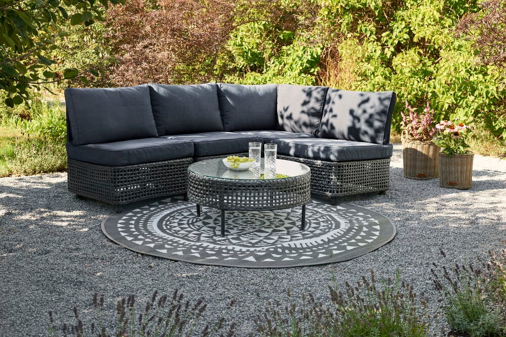 Mørkegrå utendørs hjørnesofa med Hjørnepute Split Grå/Svart, rundt glassbord og mønstret teppe.
