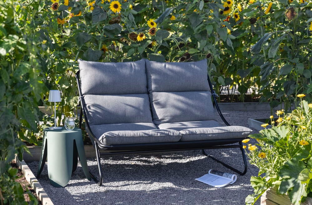 Relaxsofa Bari 135x93,5x89 cm Svart/Grå, grønt sidebord, hvit lampe, glass, magasin i en hage.