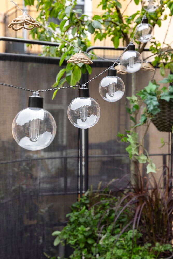 Solar-Lichterkette Stord mit klaren runden Glühbirnen, schwarzen Sockeln und geflochtenen beigen Dekorationen, in grünen Pflanzen.