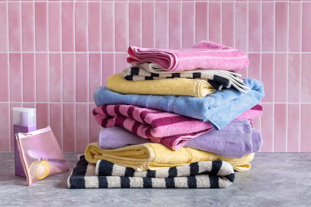 En stapel vikta färgglada Handduk Emma bomullshanddukar, en rosa väska och en lila flaska.