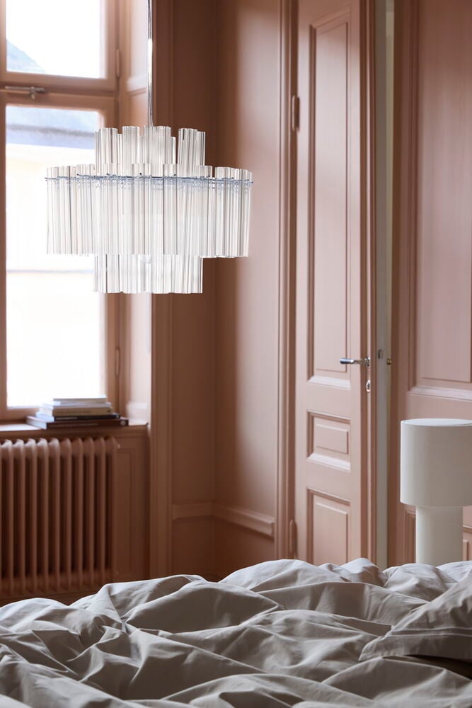Taklampa Firenze 44x34 cm, en flerskiktad ljuskrona med klara rör, med en vit bordslampa och ljusa sängkläder.