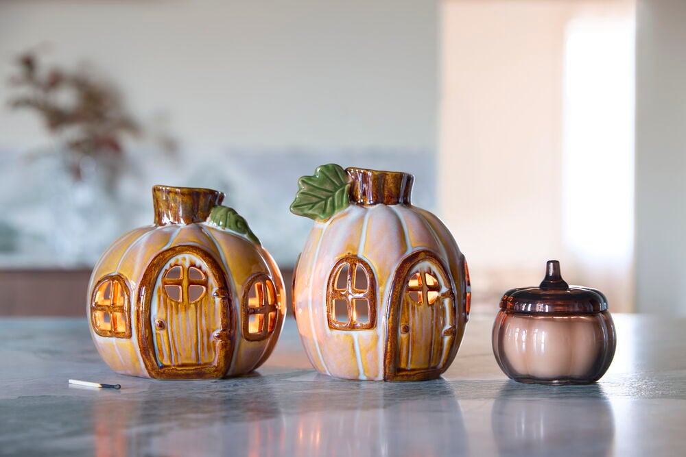 Duftkerze Pumpkin, eine Glaskerze mit dunkelbraunem Deckel, und zwei leuchtende Keramikkürbis-Kerzenhalter.