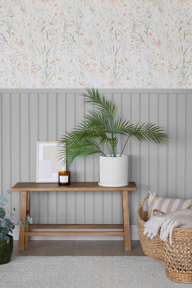 Tapete Boutique 10x0,53 m Free Flower Cream, helles Blumenmuster über grauer Paneele, mit Bank, Pflanzen und Körben.
