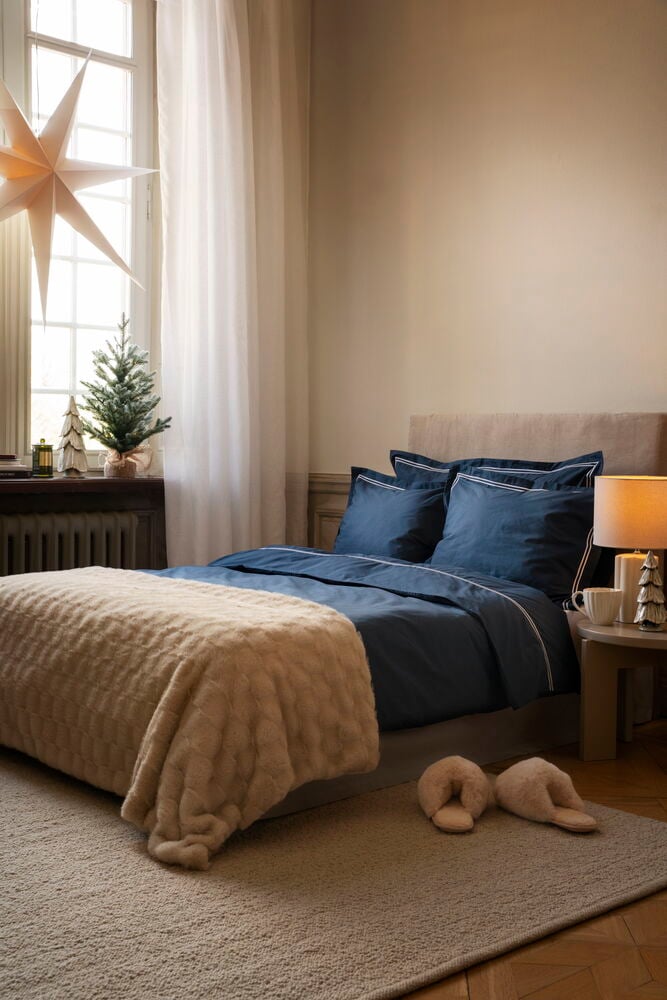 Et marineblått Sengesett Hotel Collection på en seng med beige pledd og lysbeige tøfler.