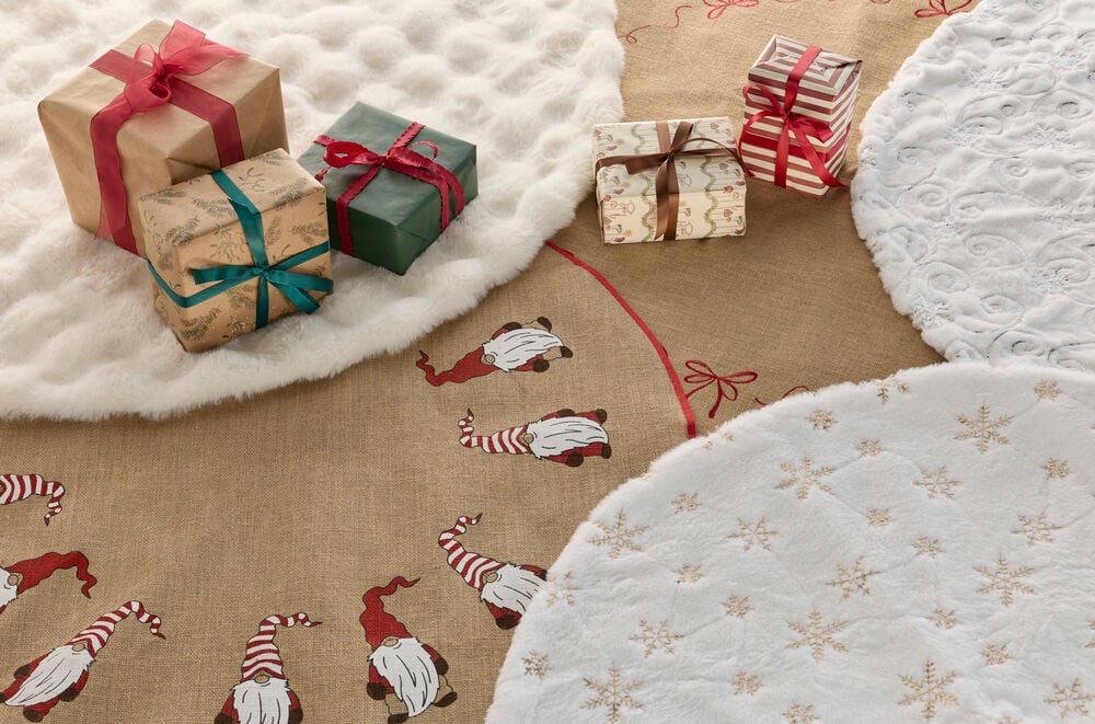 Brauner Jute-Weihnachtsbaumteppich mit roten Wichteln und Schleifendesign, weißen Plüschmatten und diverse Geschenkboxen.