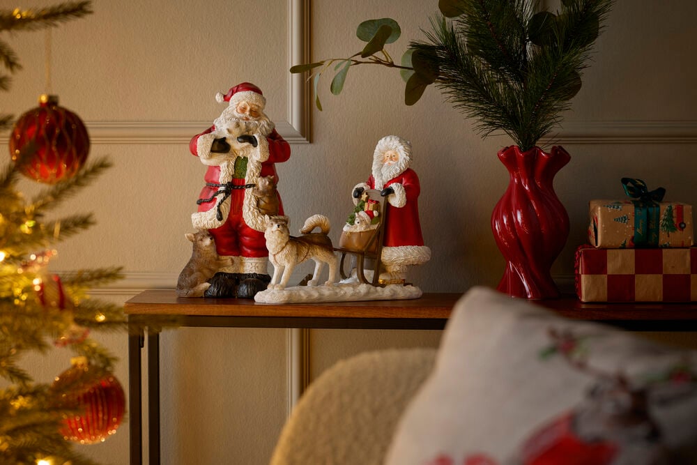 Zwei Weihnachtsmannfiguren mit Tieren und Schlitten, eine rote Vase und verpackte Geschenke.