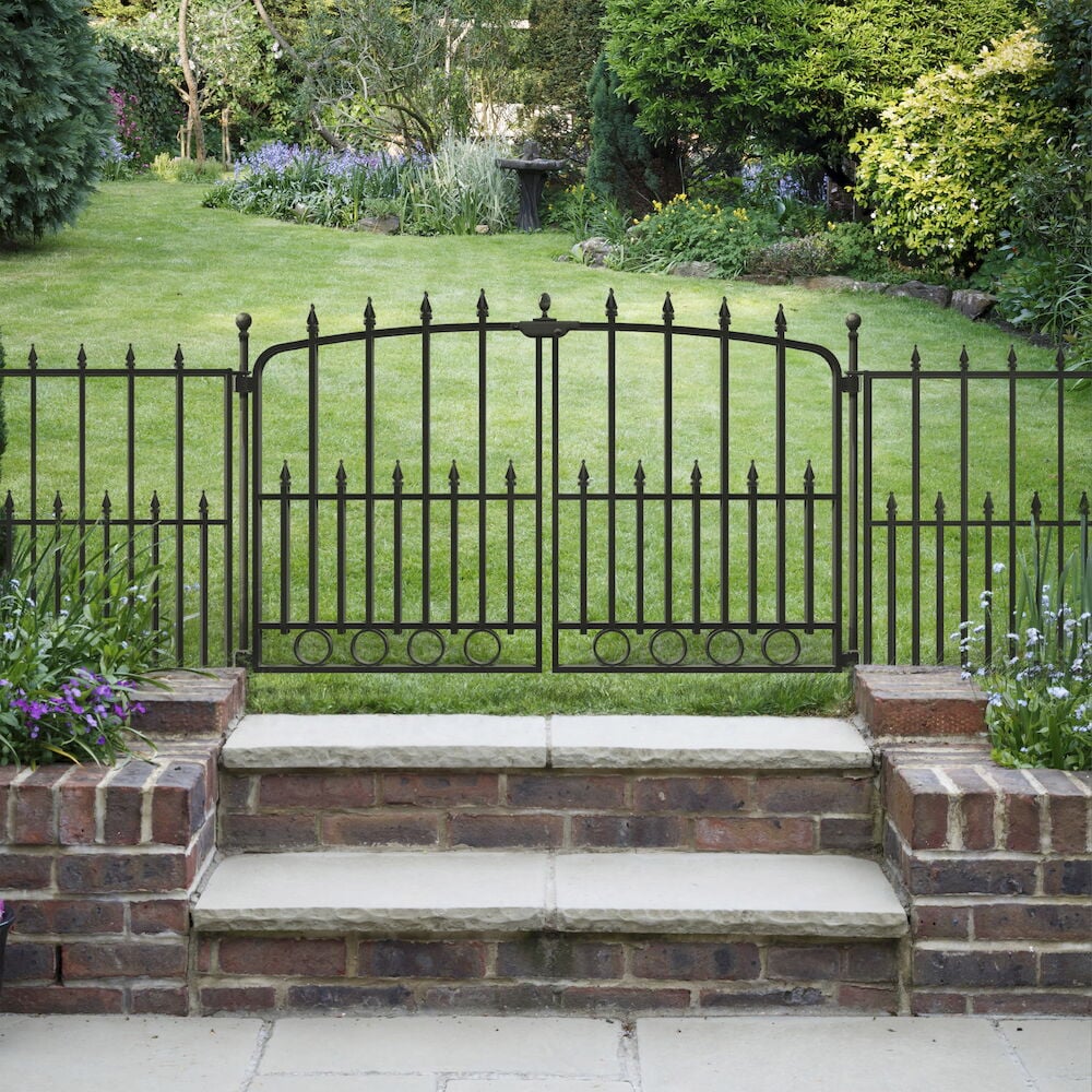 Musta metallinen portti piikkikärjillä vehreässä puutarhassa.