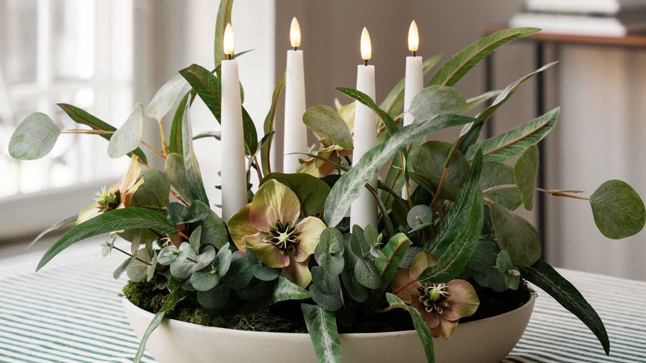 Beige skål med blomsterarrangement, grønne blader og hvite LED-lys.