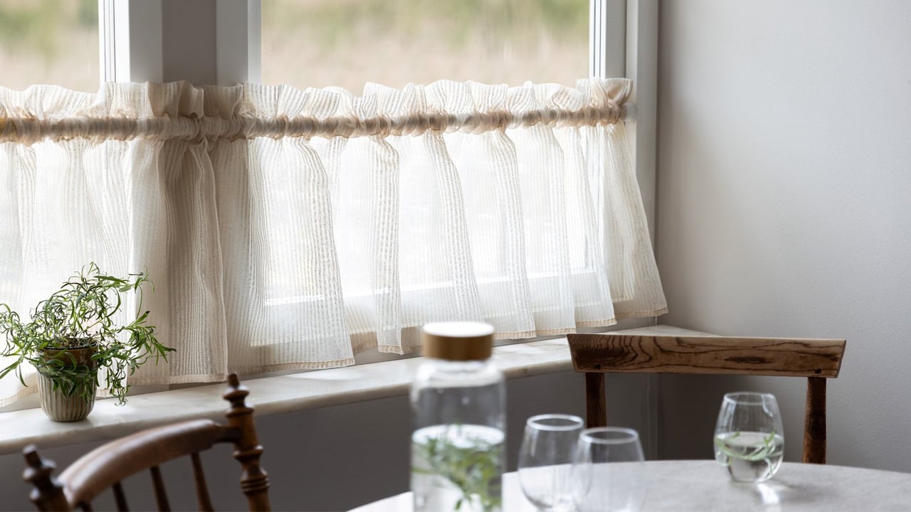 Beigefarbener transparenter Querbehang an einem Küchenfenster mit einem Teil eines runden Küchentischs im Vordergrund.