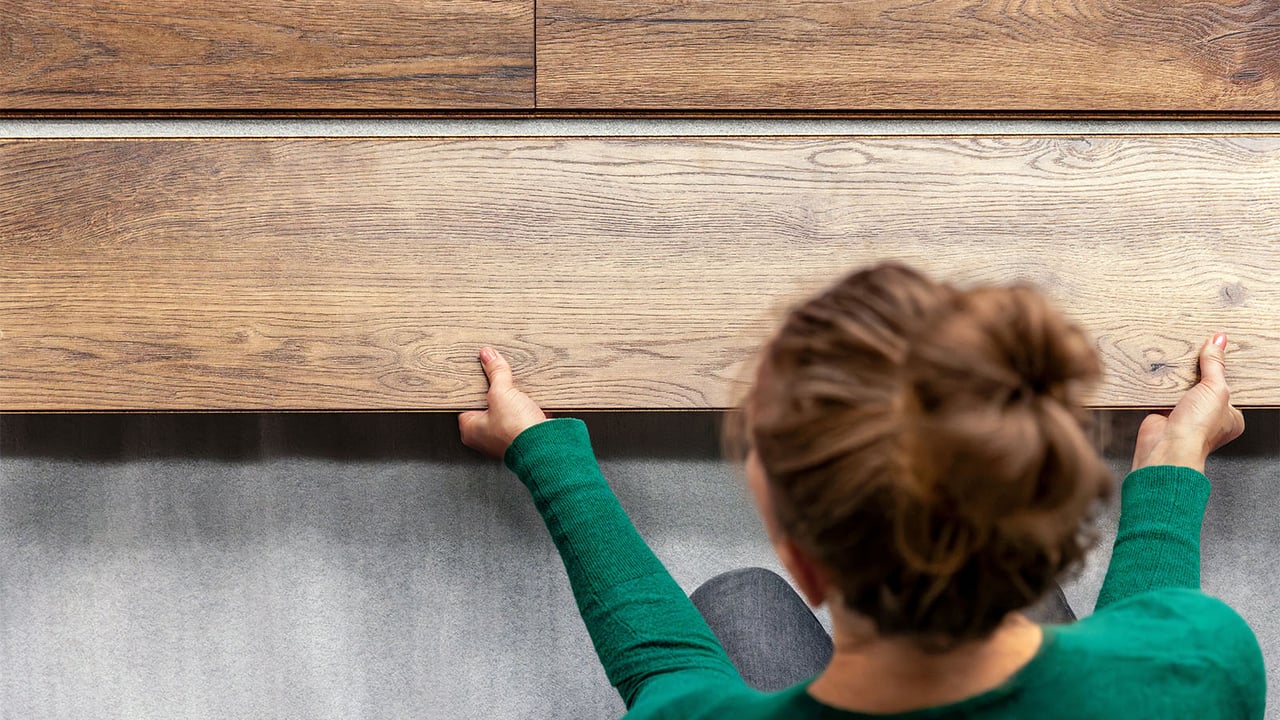 Närbild på person i grön tröja med uppsatt hår som lägger klickgolv i trä.