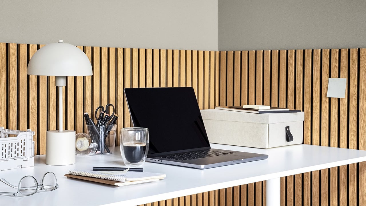 Minimalist skrivbord med laptop, lampa, kaffekopp, anteckningsblock och förvaringslåda mot akustikpanel i trä.
