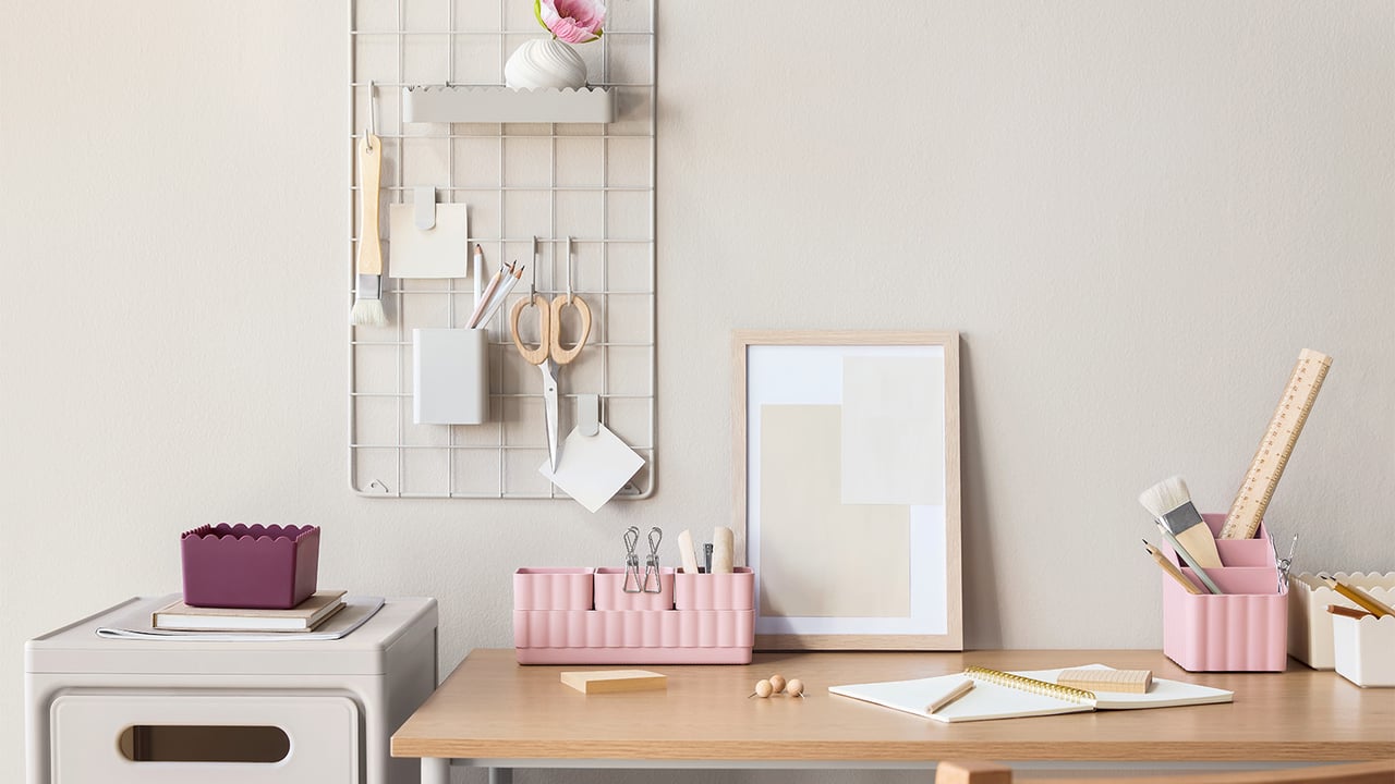 Skrivebord med rosa oppbevaringsbokser, oppslagstavle i beige metall og en beige skuffeseksjon.