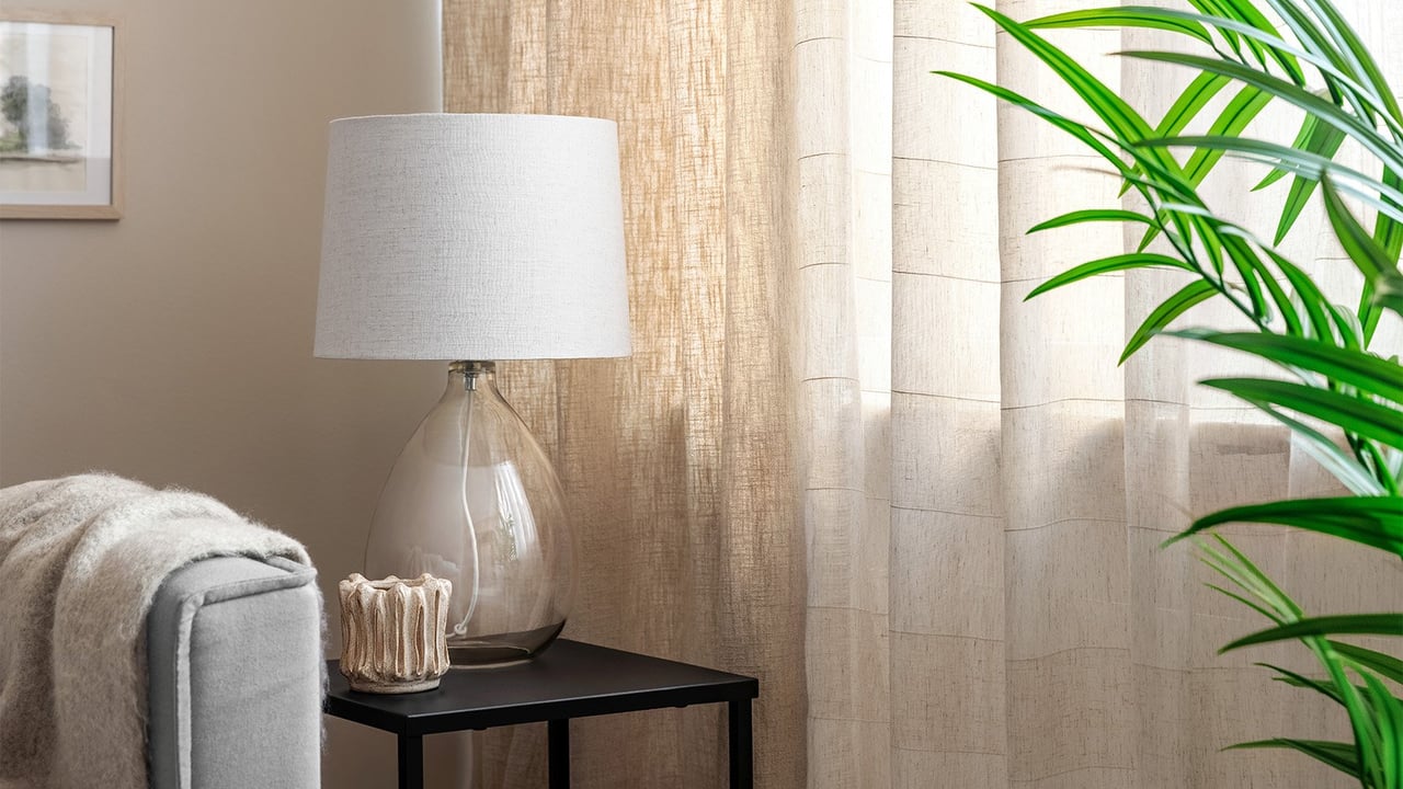Skira gardiner i naturfärger i ett vardagsrum med litet sidobord och grön växt.