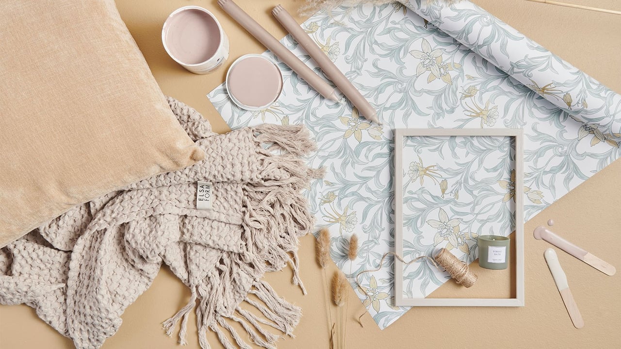 Tapetrulle, färgburk pläd och verktyg i naturnära nyanser ligger på ett golv.