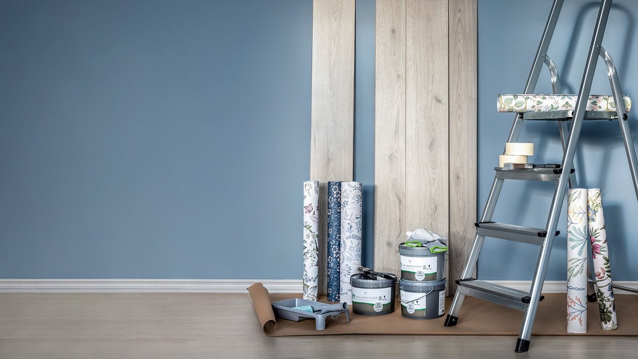 Blå vegg med gardintrapp, malingsbokser, tapet og malingsbrett i forgrunnen.