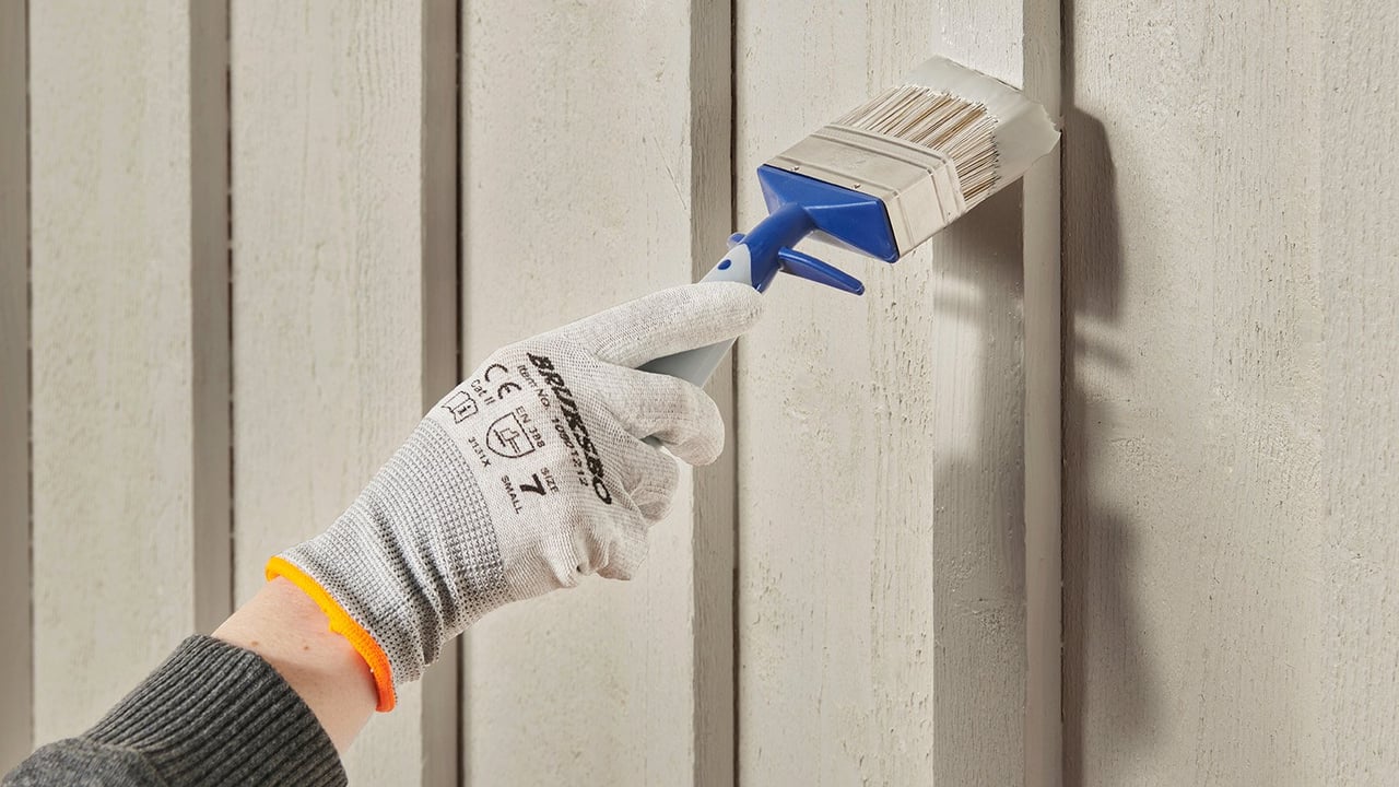 Lähikuva työkäsineellisestä kädestä, joka maalaa siveltimellä valkoista talon seinää.