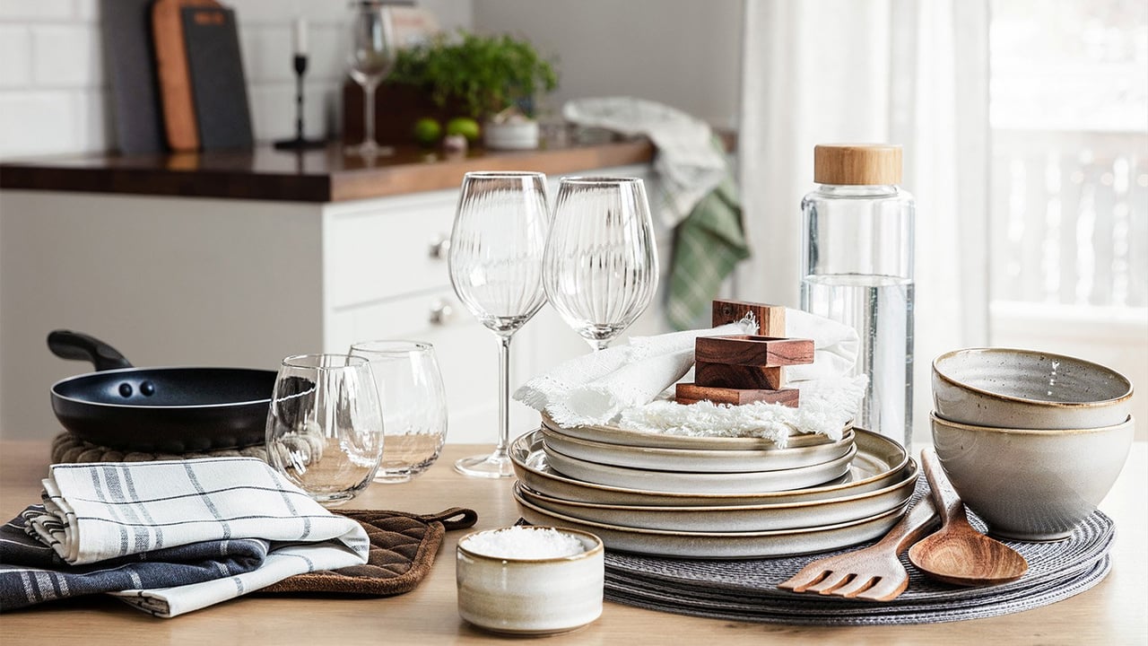 Husgeråd, porslin, stekpanna och kökstextilier uppdukat på ett bord i ett kök.