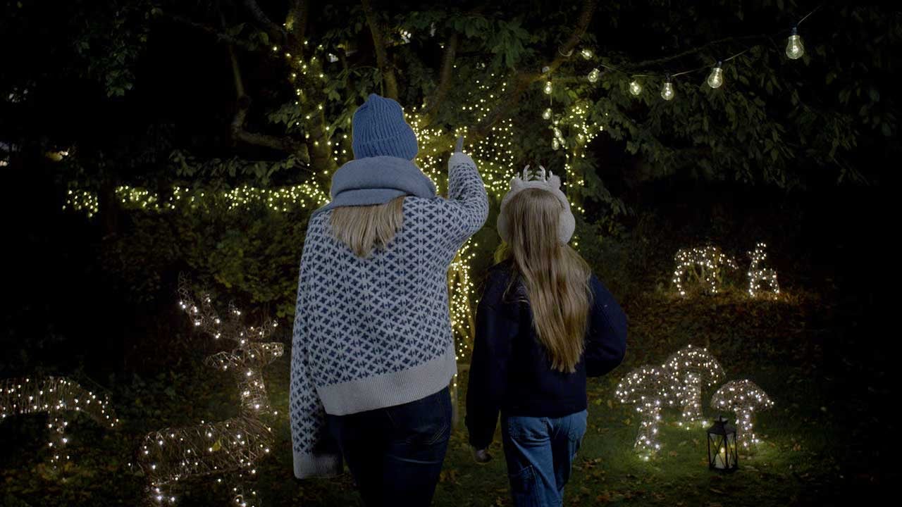 Två personer promenerar genom en mörk trädgård upplyst av ljusslingor och dekorationsbelysning.