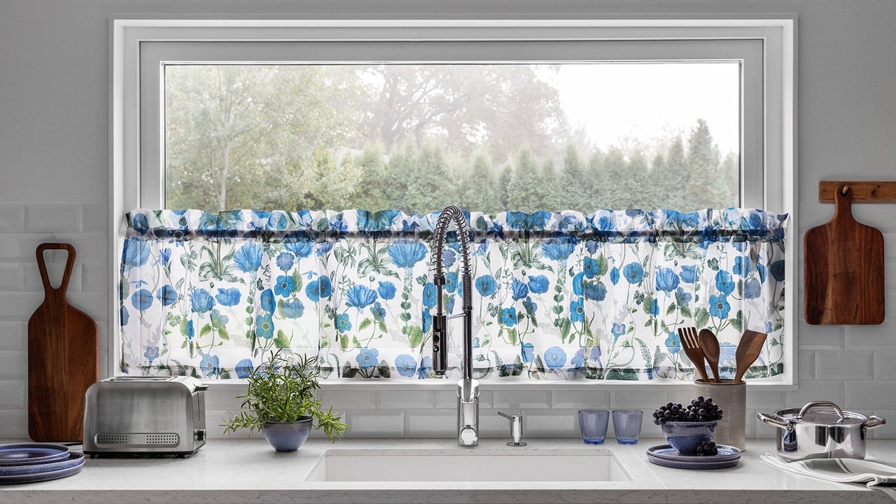 Küchenarbeitsplatte mit Spüle und Küchenaccessoires vor einem Fenster mit blauem Querbehang mit Blumenmuster.
