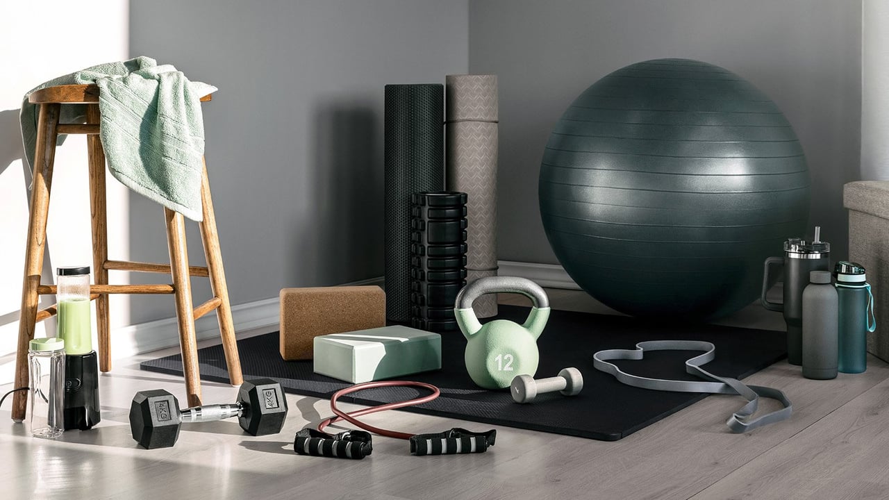 Kotikuntosali, jossa on pilatespallo, joogamatto, vastuskuminauhat ja käsipainot harmaaseinäisessä huoneessa.