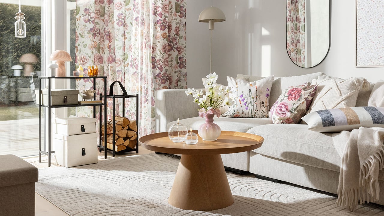 Vardagsrum med soffa, soffbord och matta i ljusa toner med blommig gardin och soffkuddar.