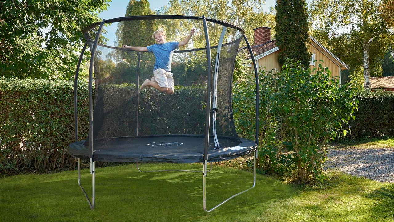 Pojke i blå t-shirt och shorts hoppar på en studsmatta som står i en grönskande trädgård.