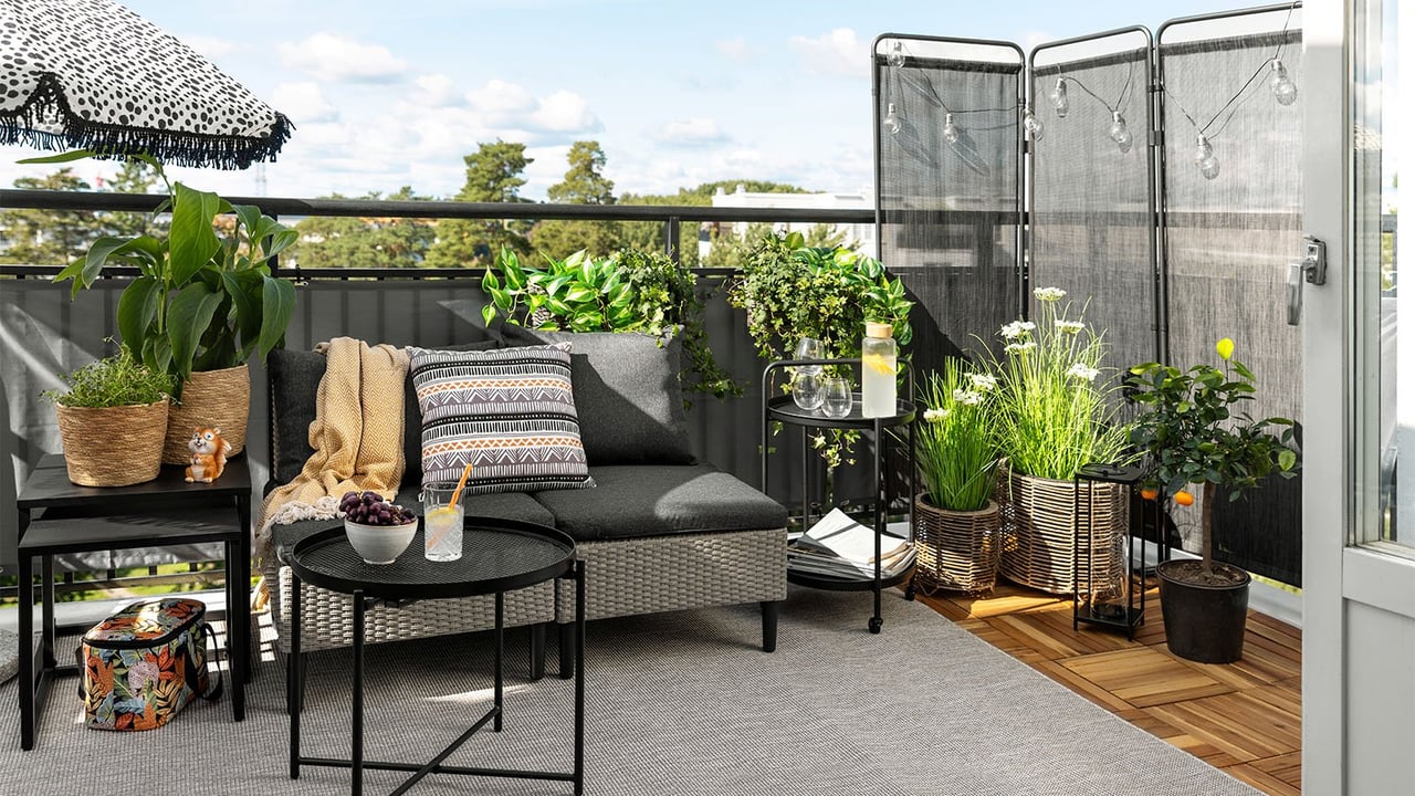 Großer Balkon mit Sichtschutz und Lounge-Bereich, Pflanzen in Körben und Blumentöpfen und einem gepunkteten Sonnenschirm.