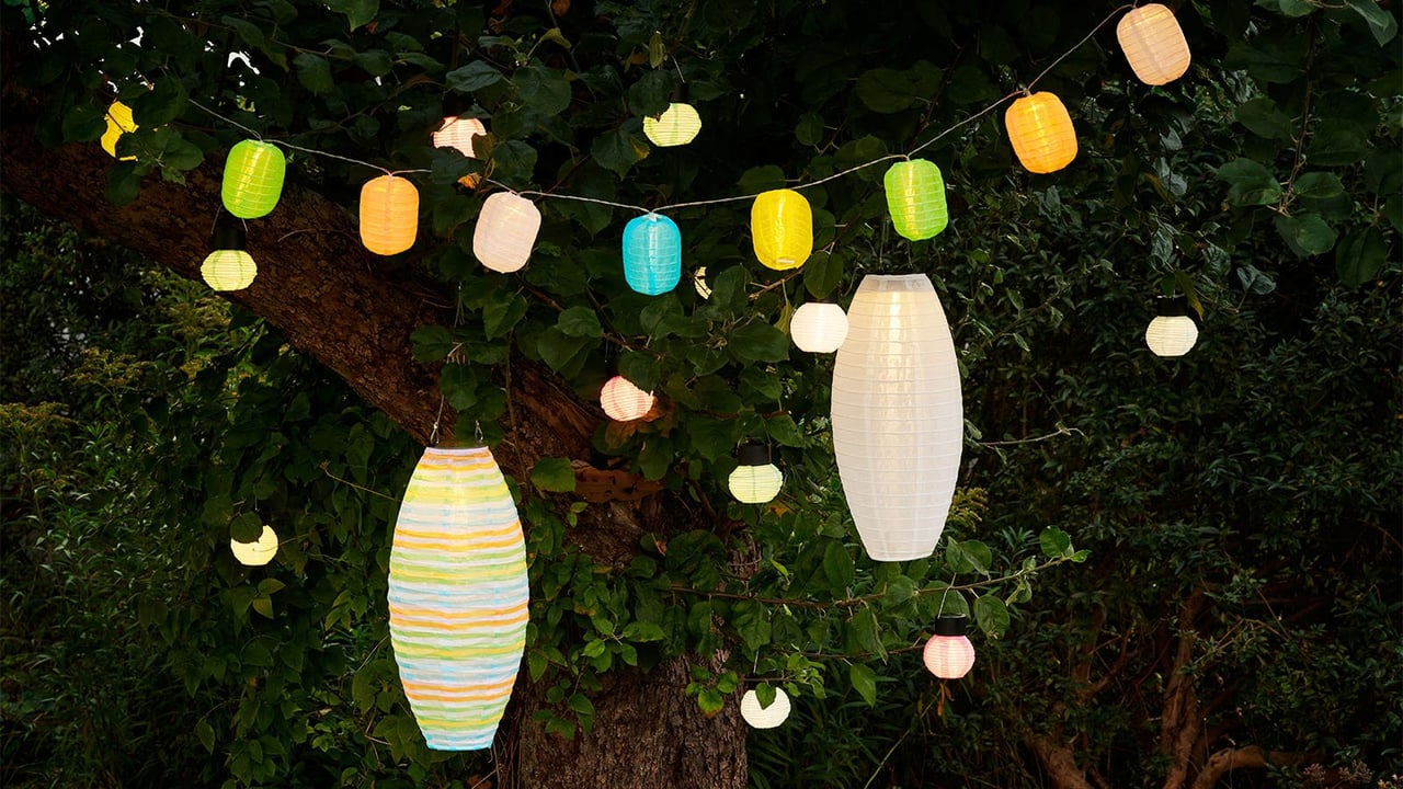 Yksittäisiä valkoisia riisipaperivalaisimia ja ketjuja, joissa on värikkäitä pieniä riisipaperivalaisimia riippumassa puissa pimeässä.