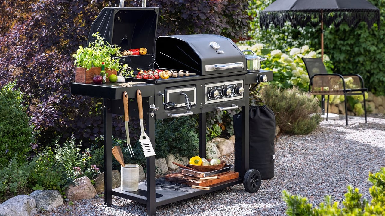 Kohle- und Gasgrill mit Grillutensilien auf einem Kiesweg im Garten.