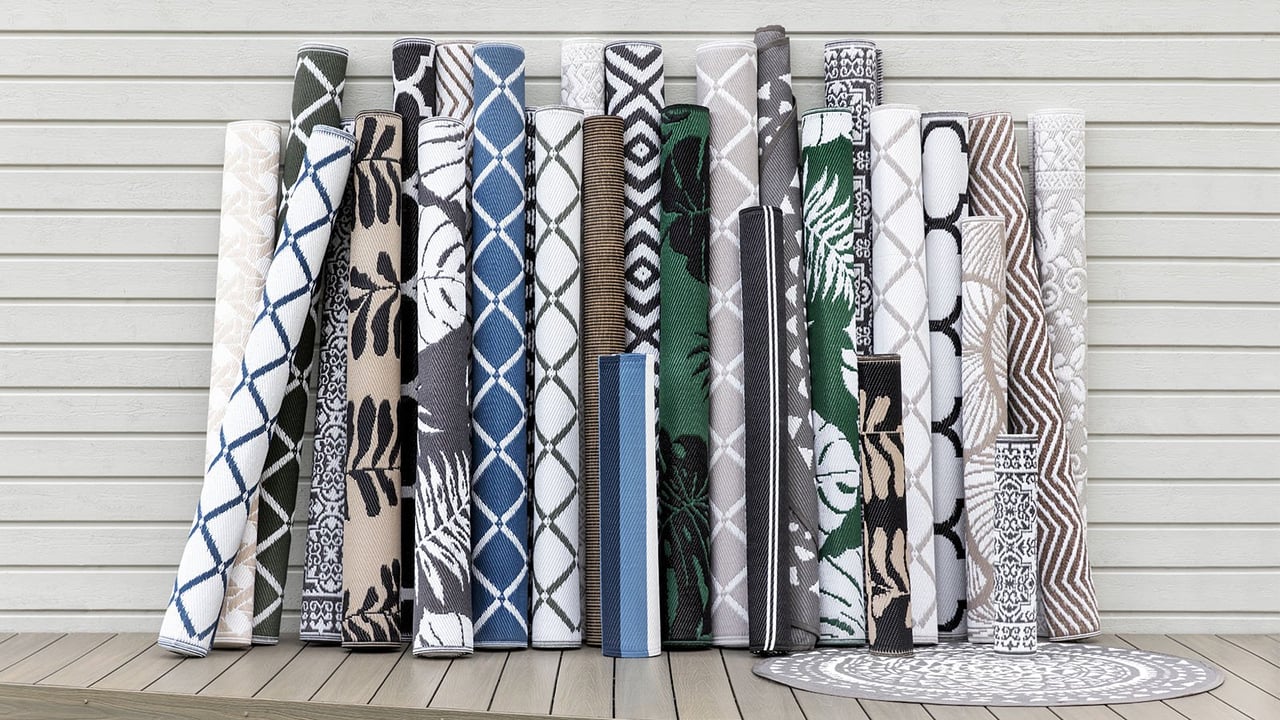 Flera ihoprullade utomhusmattor i olika storlekar och mönster lutandes mot ett vitmålat plank.