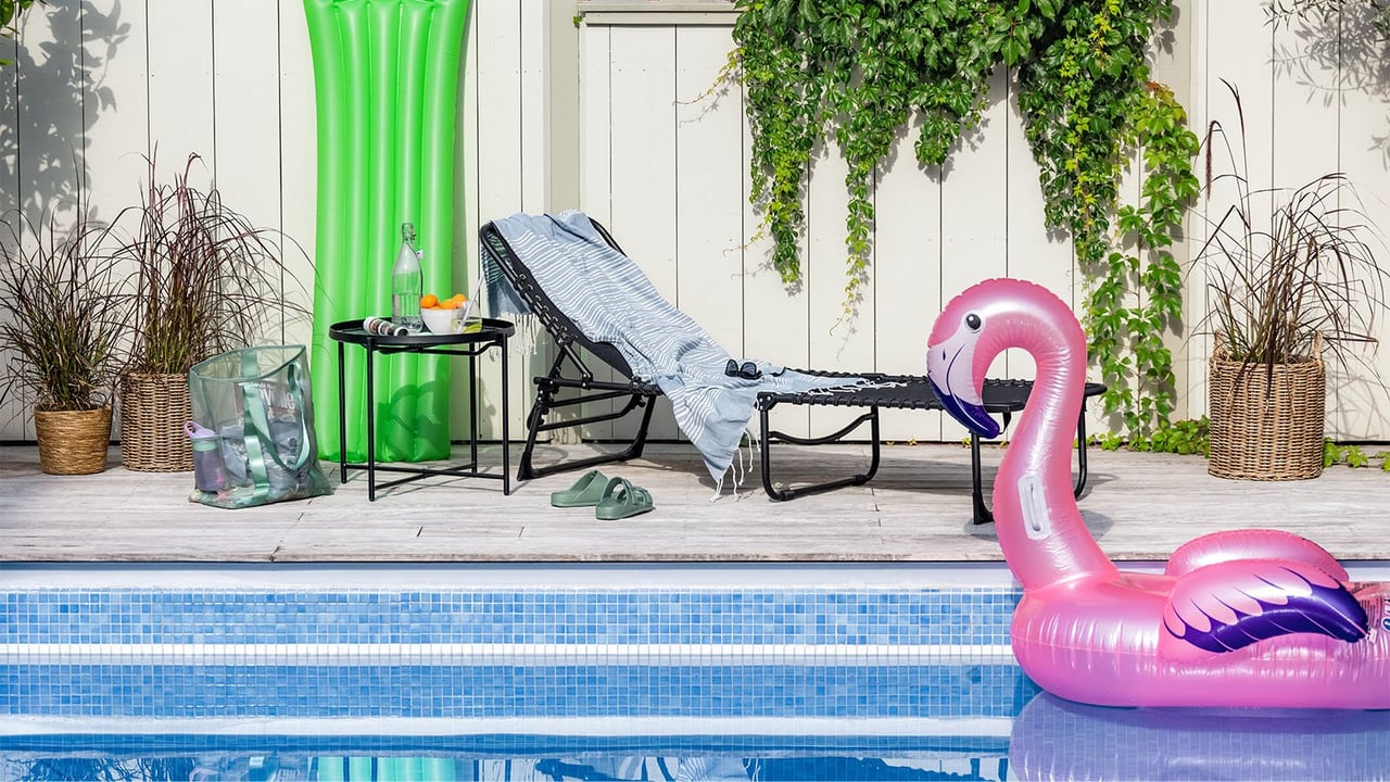 Uima-allas, jossa on puhallettava vaaleanpunainen flamingo. Uima-altaan vieressä on aurinkovuode, pieni pöytä ja muita vesileluja.