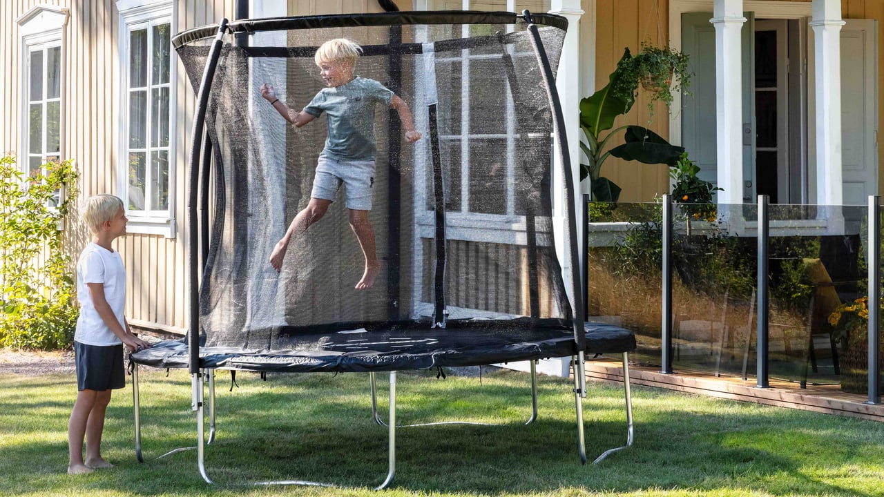 Lapset hyppivät turvaverkolla varustetulla trampoliinilla pihalla keltaisen talon edessä.