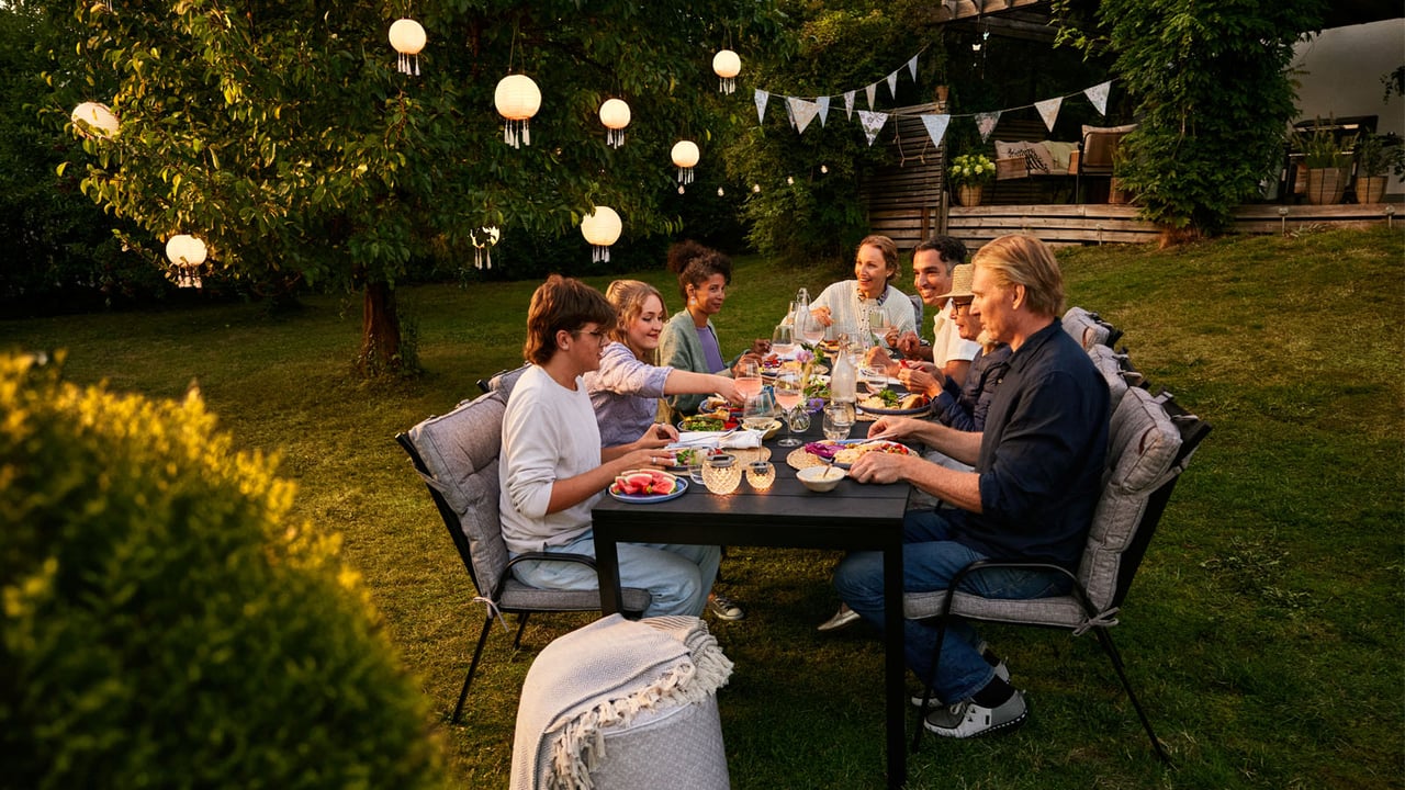 En sommerlig hagefest i kveldslys der venner samles rundt et langbord med mat, lyslykter, vimpler og solcellebelysning i treet.