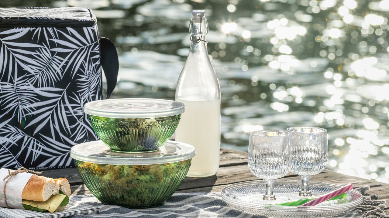 Picknick mit Nudelsalat in runden Lunchboxen, Gläsern, Tellern und einer Kühltasche auf einem Steg, umgeben von glitzerndem Wasser.
