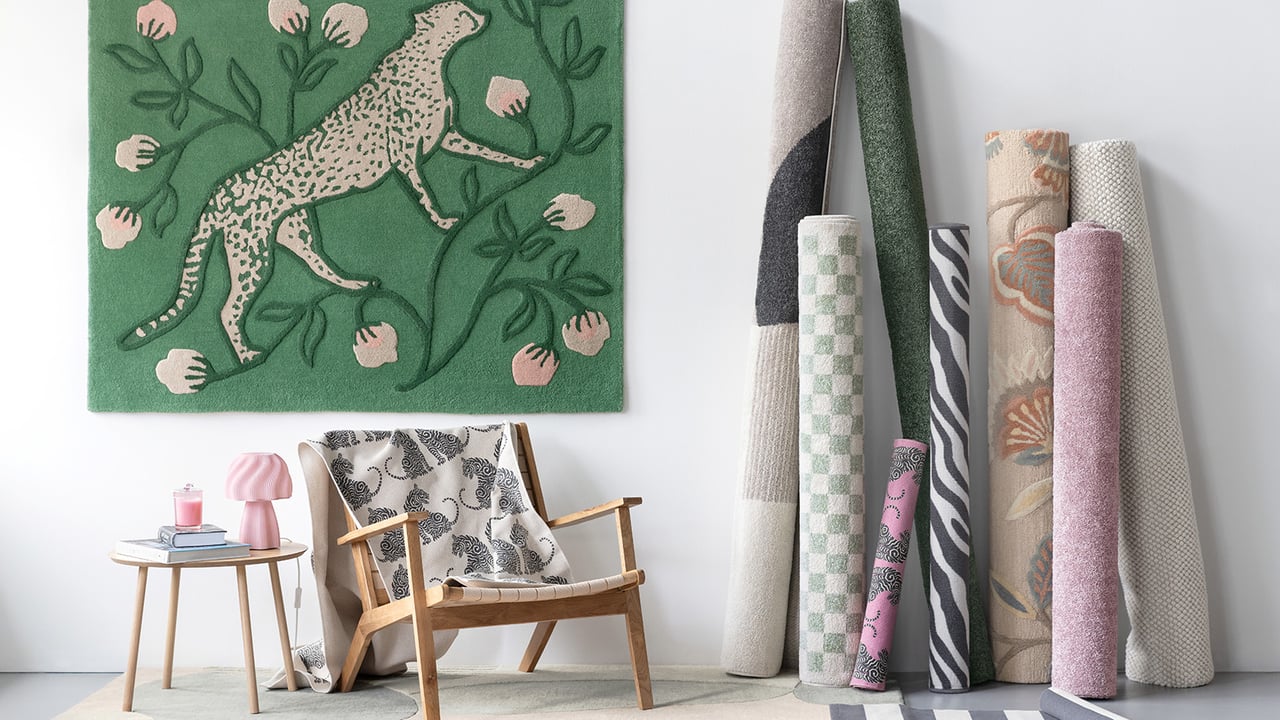 Zusammengerollte Teppiche in verschiedenen Mustern und Farben, ein rosa Rollwagen und ein hängender grün karierter Teppich.