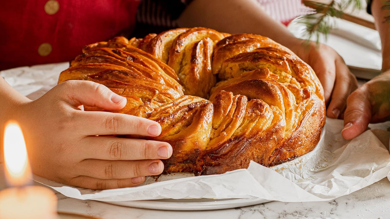 Hände, die einen frisch gebackenen Safrankranz auf einem Teller befühlen. Daneben eine brennende Kerze.